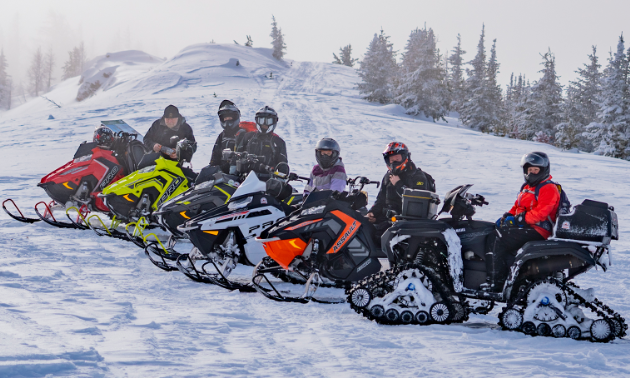A row of snowmobile riders pose for a photo on a snowy hill. 
