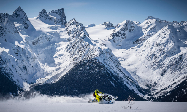 A snowmobiler carves tracks in a field in front of massive mountains.