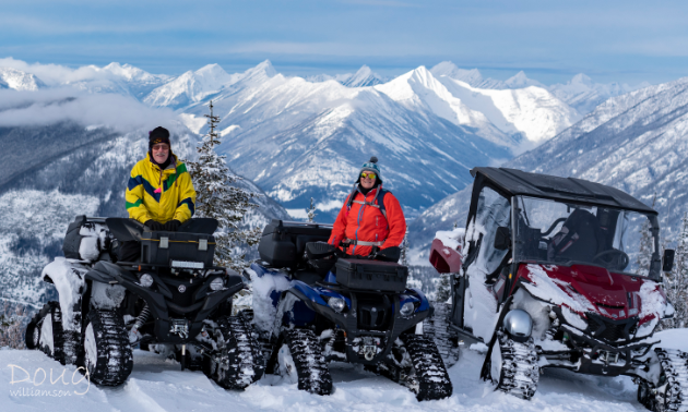 An elderly couple sit on their ATVs with tracks on top of a snowy mountain.