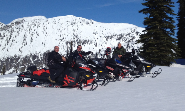 A row of snowmobilers smile on their sleds with a mountain in the background.