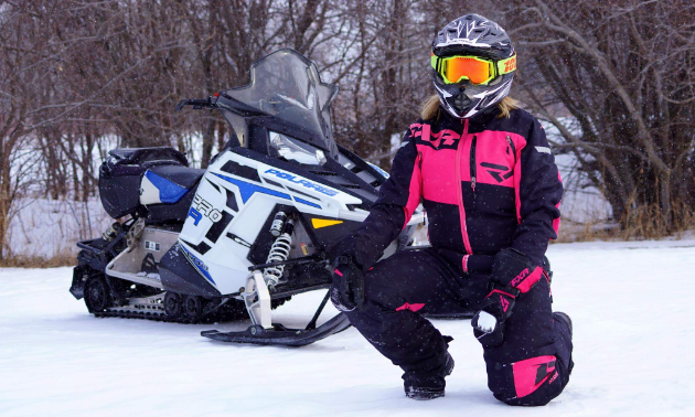 A woman wearing pink and black clothes kneels in front of a white and black and blue snowmobile in Gimli, Manitoba.