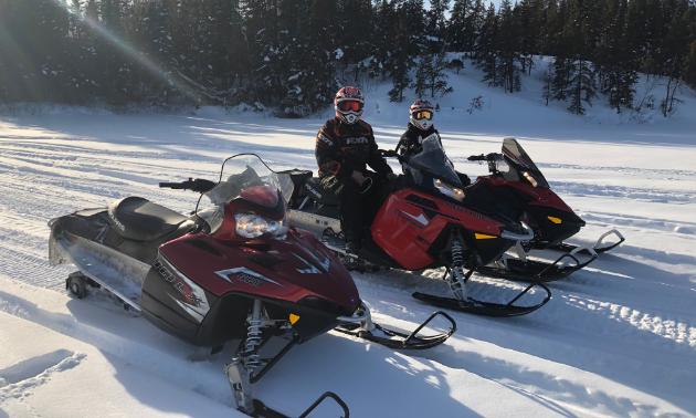 Three snowmobiles sit in a line with two occupied by sledders.