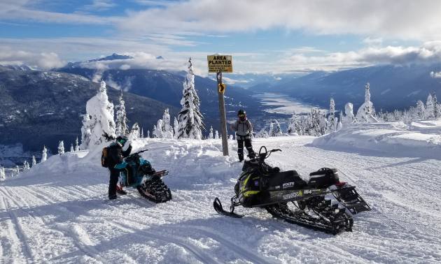 A couple riders stand next to snowmobiles at a summit in Revelstoke.