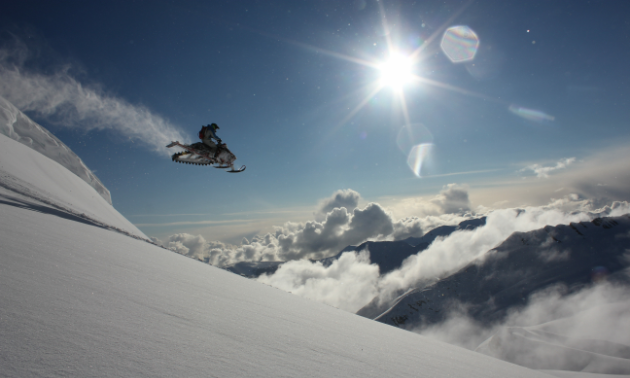 A snowmobiler gets massive air off a mountain as the sun shines overhead.