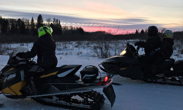 Two snowmobilers pause to look at a pink sunset.