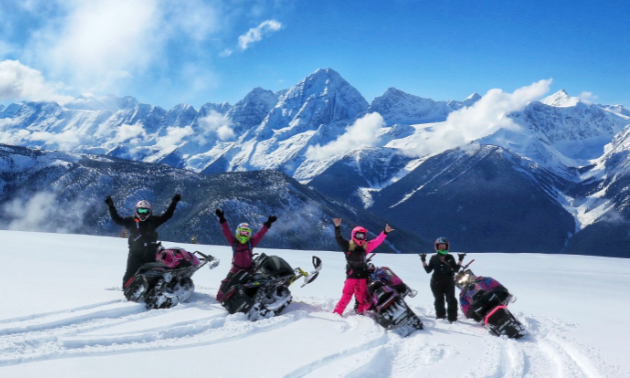 Four women hold their arms up as they stand next to their snowmobiles in front of the Rocky Mountains.