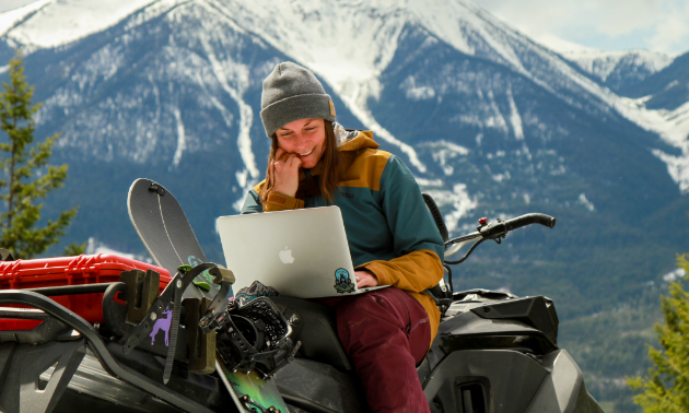 Brittney Dickson sits on her snowmobile while looking at her laptop in the mountains.