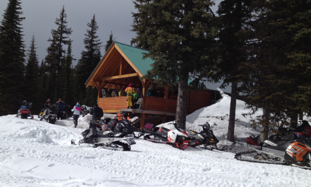 Ingersol Cabin is wooden, large and has a green roof. An assemblage of snowmobiles are parked up front.