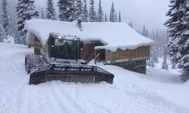 A groomer plows snow in front of a cabin.