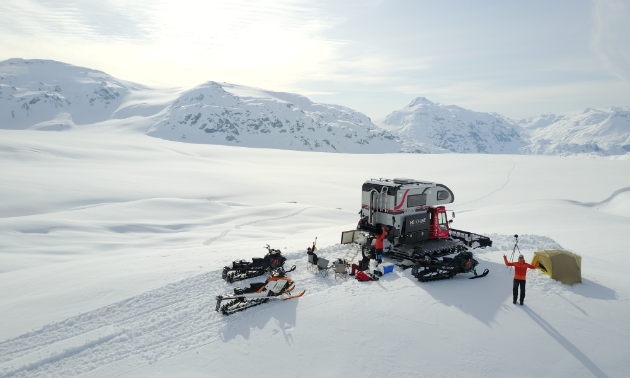 A groomer and two snowmobiles are parked on a snowy, isolated plateau.