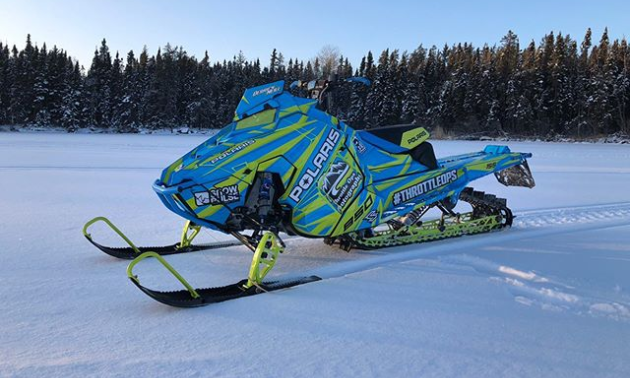 A blue and neon green snowmobile on the snow with the woods in the background.