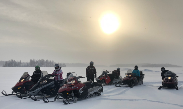 Six snowmobilers stop on a flat snowy field near Flin Flon Manitoba with the sun trying to shine through the clouds in the background.