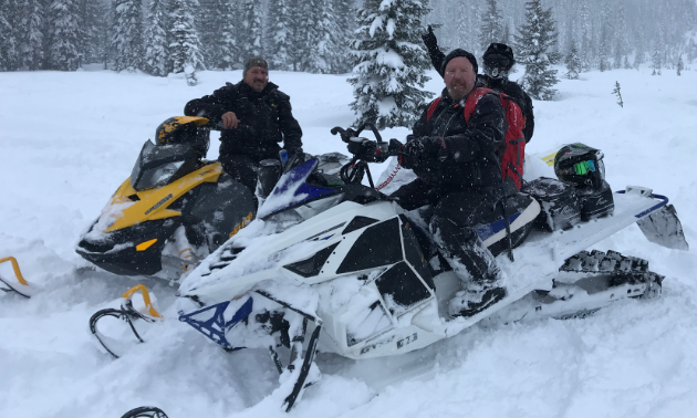 Three snowmobilers pose in a meadow as snow falls around them.