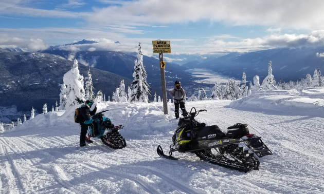 Two snowmobilers park their snowmobiles for a picture on Frisby Ridge with Revelstoke in the background.