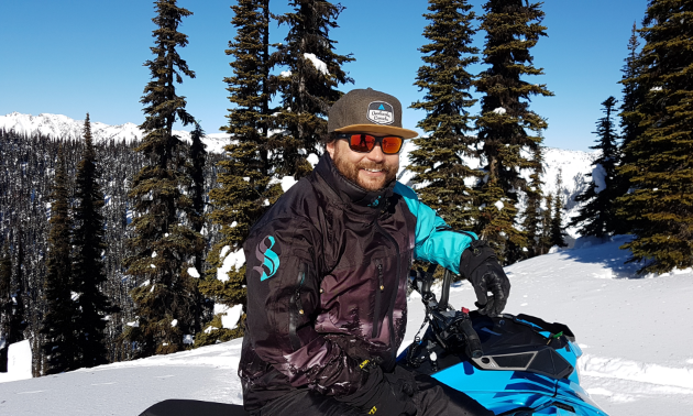 Curtis Pawliuk smiles on his blue snowmobile on a snowy, tree-filled hill.