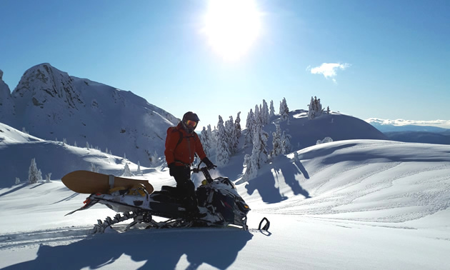A man stands on a snowmobile with the sun hovering overhead. 
