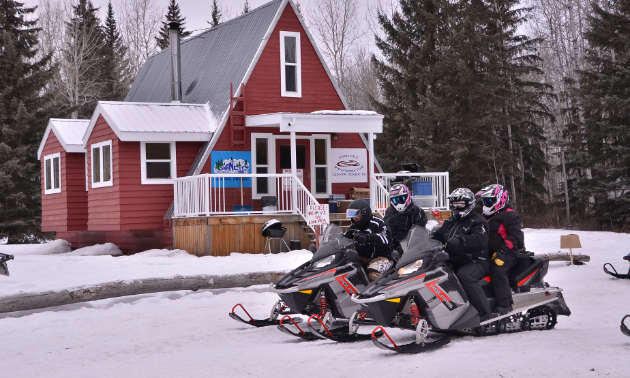 Two snowmobiles carrying three riders prepare to embark on adventure in front of a red cabin in the woods. 
