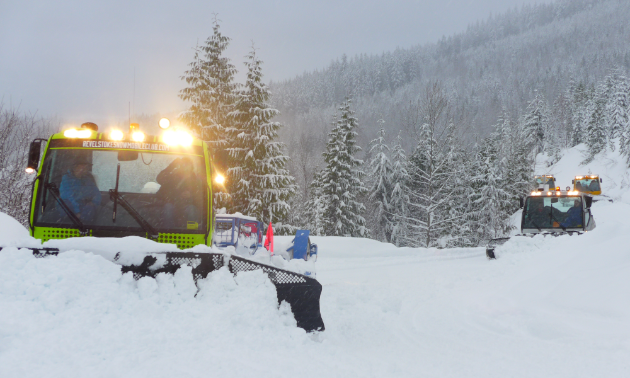 Several snowcat groomers plow trails on a snowy mountain.