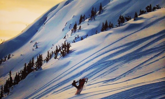 A snowmobiler pops a wheelie on a steep hill as the sun sets in the purple distance beyond the mountain.