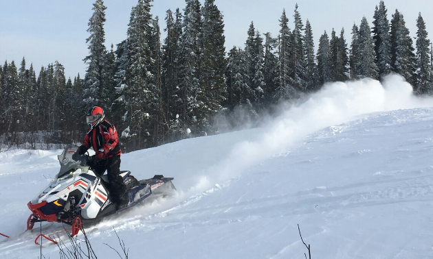 A plume of white smoke puffs up from a snowmobile as it makes its way down a hill.