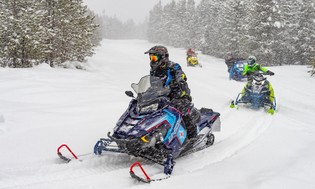 A line of snowmobiles ride through the snow. 