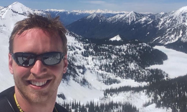 Josh Salzmann has short brown hair and is smiling while wearing sunglasses. The South Purcell Mountains are in the background.