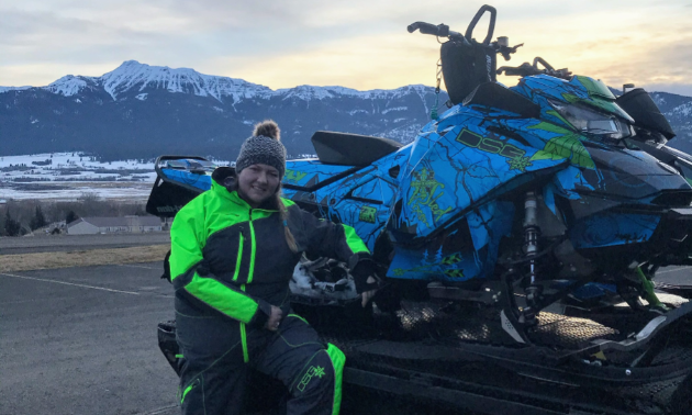 Kadie McCallum wears a green and black coat and poses next to her blue and green snowmobile parked on a trailer.