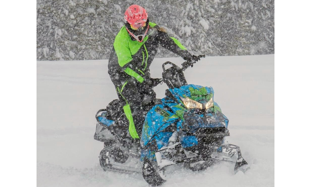 Kadie McCallum wears a pink helmet with a green and black coat and rides a blue and green snowmobile through a snowstorm.