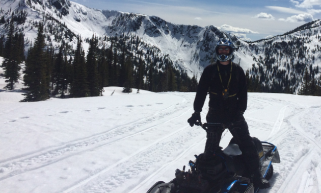 A snowmobiler stands on his ride on a snowmobile trail with mountains in the background.