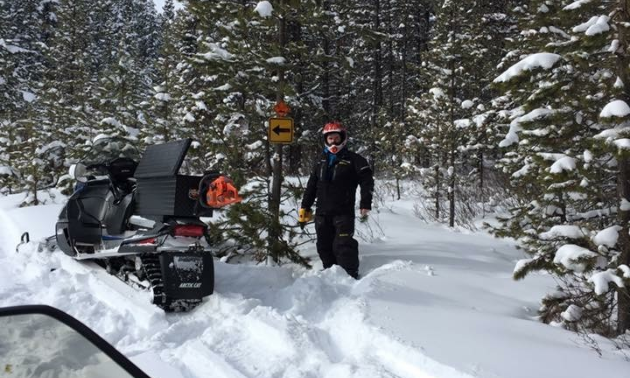 A man stands next to a sign and his parked snowmobile on a snowmobile trail.