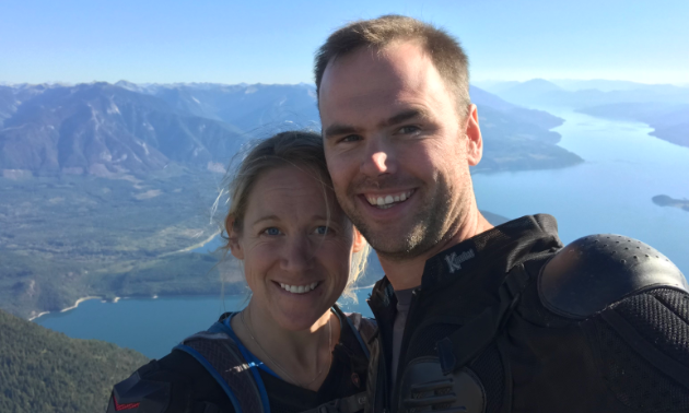 Matt Elliott and Sarah Blancher smile from a high vantage overlooking a lake and mountains.