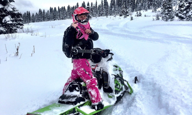 Meghan Bosecker wears pink and black riding gear while standing on her snowmobile on a trail.