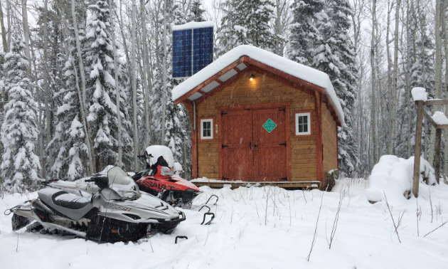 A wooden shelter with two snowmobiles parked up front.