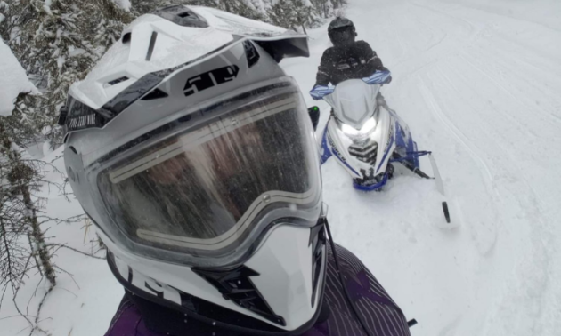 A snowmobiler looks into the camera from close range on a trail among trees.
