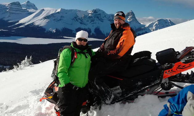 Les and Amanda L’Heureux smile on their snowmobiles with mountains in the background.