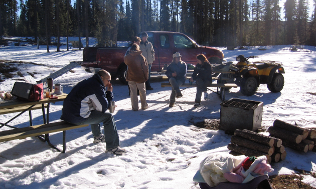 A family takes a break at picnic tables in the snow. A truck and ATV are parked nearby. 

