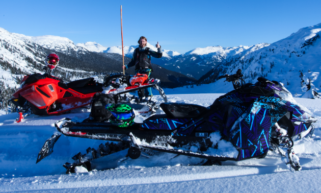 Ryan Thorley stands next to an orange bamboo pole with two snowmobiles in the foreground and snow-capped mountains in the background.