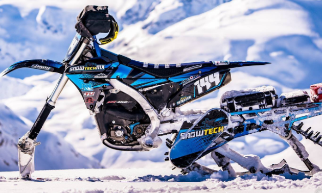 A side profile of a blue and white snow bike with snow-covered mountains in the distance.