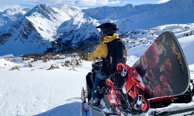 A snowmobiler looks off into the distant mountains. They have a red and black snowboard on their sled.