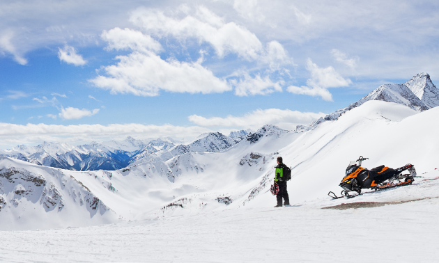 A snowmobiler gets off his sled to take in a view of the mountains.