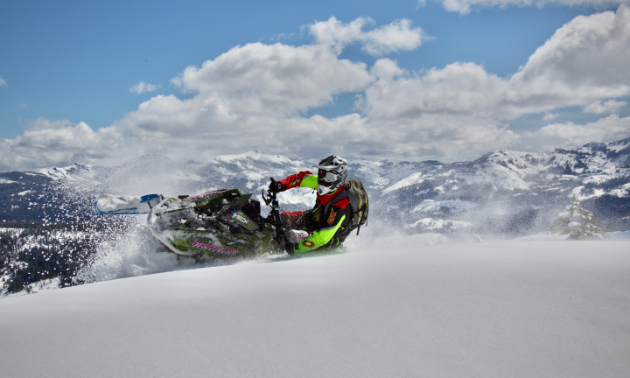 A snowmobiler banks into fresh snow while driving towards the camera. 