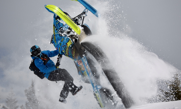 A snowmobiler rides his snowmobile upside down with only his hands on the handlebars. 