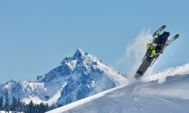 A snowmobiler is suspended in the air at the top of a mountain with more snow covered peaks in the distance.