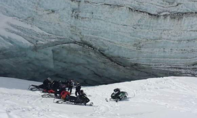 A group of snowmobilers gather under a mountain near a large snowpack. 
