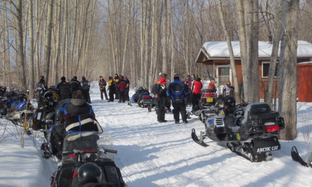 A row of snowmobiles are gathered to take while riders socialize in the background