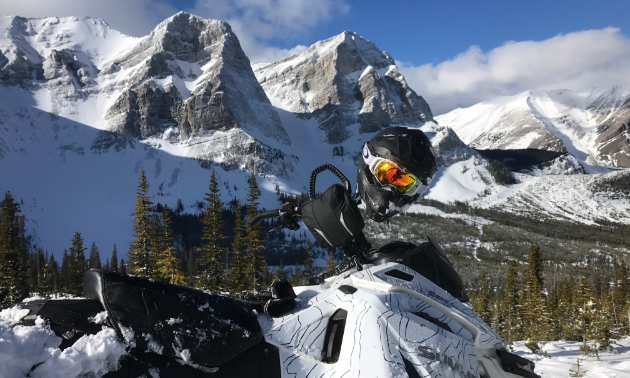 A snowmobile has a helmet on the handlebars in the foreground and towering mountain peaks in the background. 
