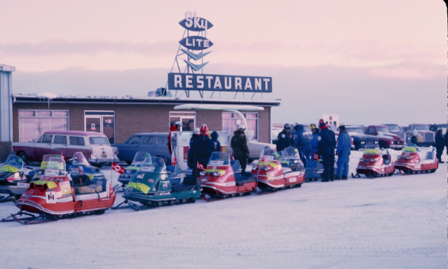 A picture of a line-up of snowmobiles parked outside a diner on a winter day. 