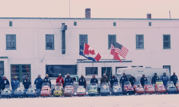 Snowmobile riders line up for the Manitoba Centennial in front of a building