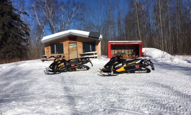 A warm-up shelter with a shed beside it and two snowmobiles parked in front in Swan River.