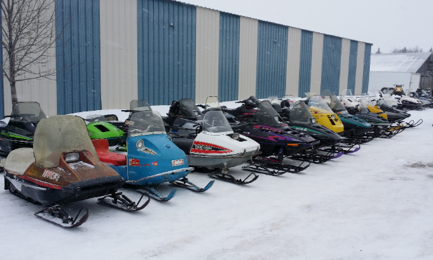 A row of vintage snowmobiles is parked beside a building